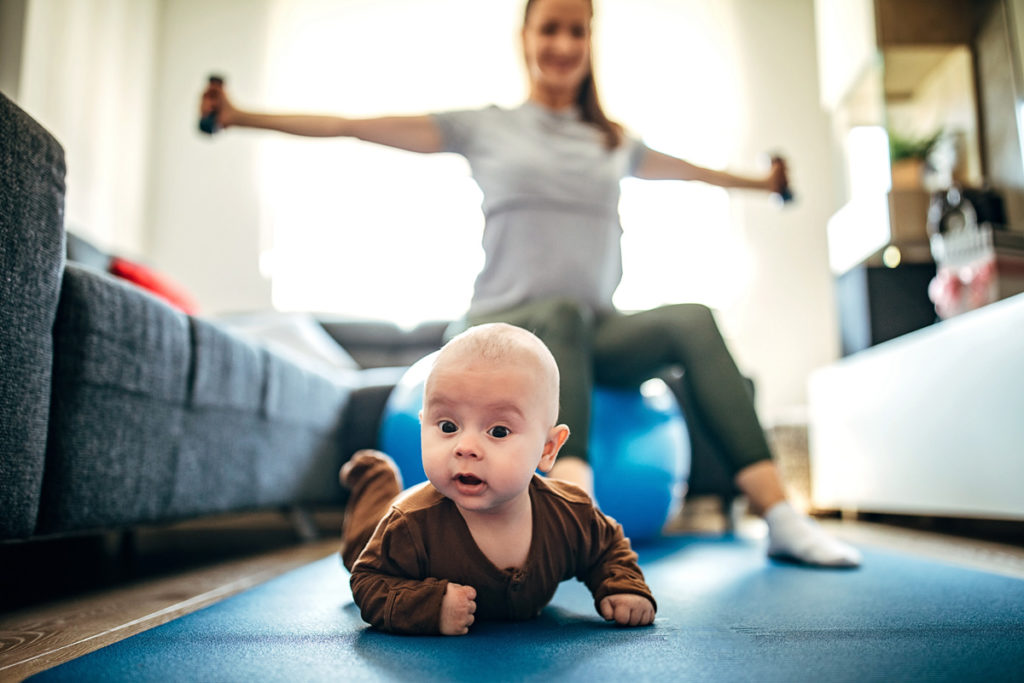 Baby crawling in front of mother working out with dumbells