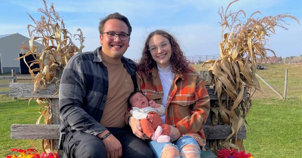 Mom, dad and baby sit outside on a fall day smiling.