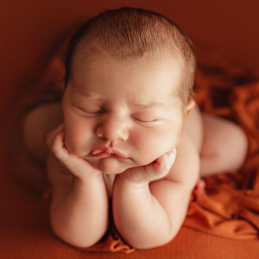 baby laying in orange blankets with hands perched on chin.