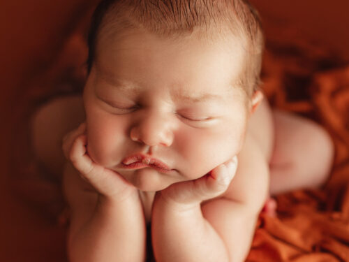 baby laying in orange blankets with hands perched on chin.